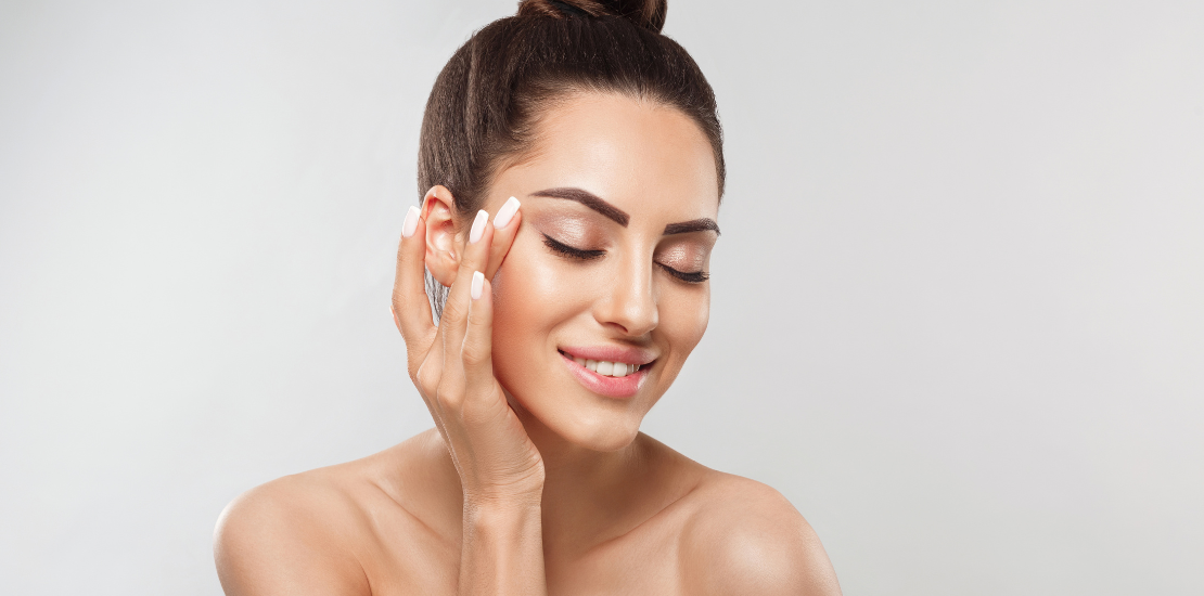 Seorang wanita yang tampak puas dengan gaya rambut sanggul lembut menyentuh sisi wajahnya, menunjukkan kulit yang halus dan bersih dengan tampilan makeup yang halus