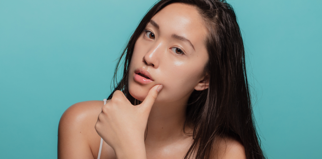 Seorang wanita muda dengan rambut hitam panjang, duduk dengan pose memegang dagu, menampilkan wajah yang cerah dan berkilau dengan latar belakang biru